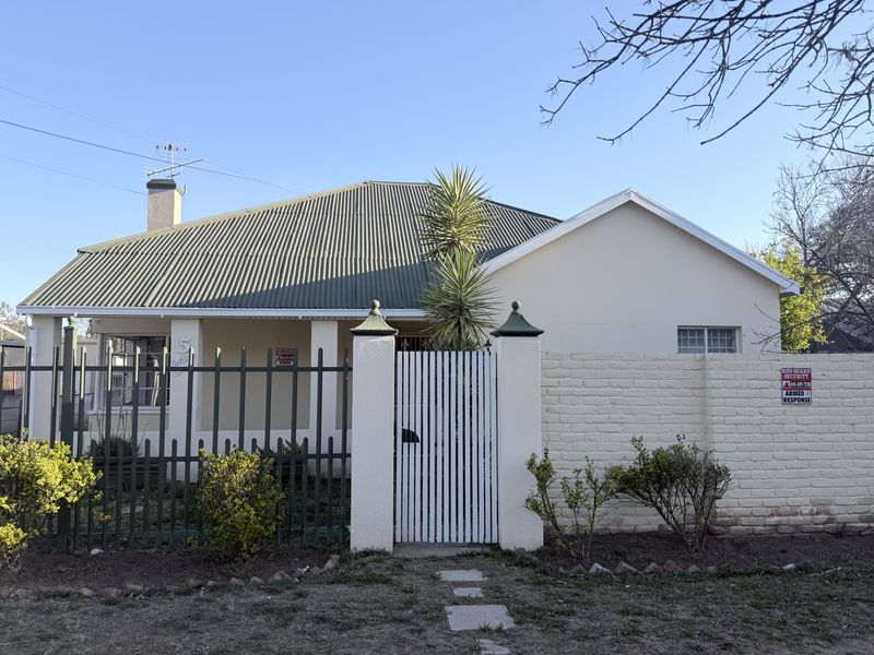 Front elevation of a single-story house featuring a corrugated metal roof, covered porch, and secure perimeter fencing with a pedestrian gate.