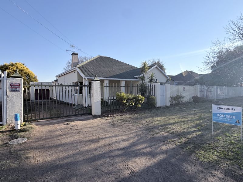 Front view of a single-story house featuring a corrugated iron roof, chimney, and secure metal gate entrance with a gravel driveway.