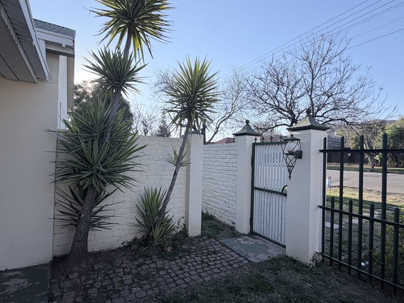 Front entrance area featuring a painted brick boundary wall, secure pedestrian gate, wrought iron fencing, and cobblestone paving with established landscaping.