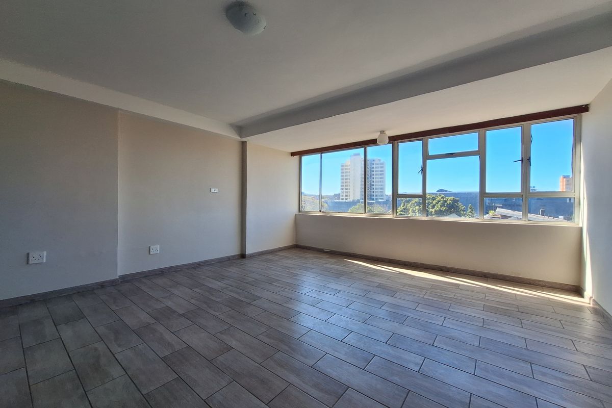 A bright and spacious empty living area featuring modern wood-look tiled flooring and large windows that provide ample natural light and urban views.