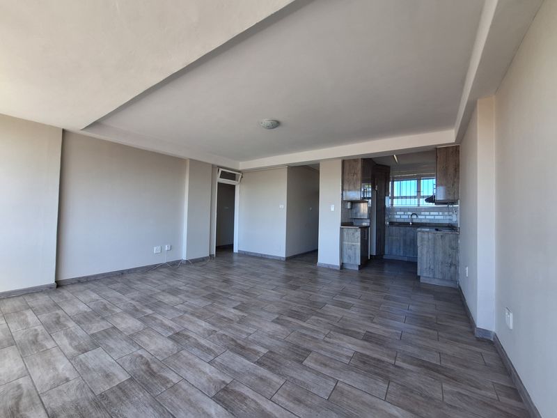 Spacious open-plan living area featuring modern wood-look floor tiles, a recessed ceiling, and a compact, functional kitchen with built-in cabinetry.