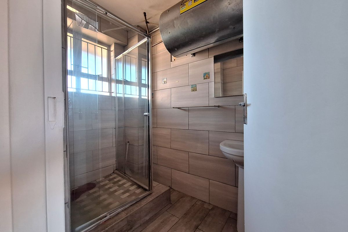 A functional bathroom featuring a shower enclosure, pedestal basin, and tiled finishes, with an exposed geyser unit.