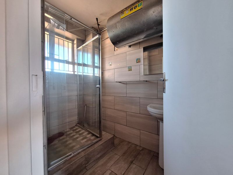 A functional bathroom featuring a shower enclosure, pedestal basin, and tiled finishes, with an exposed geyser unit.