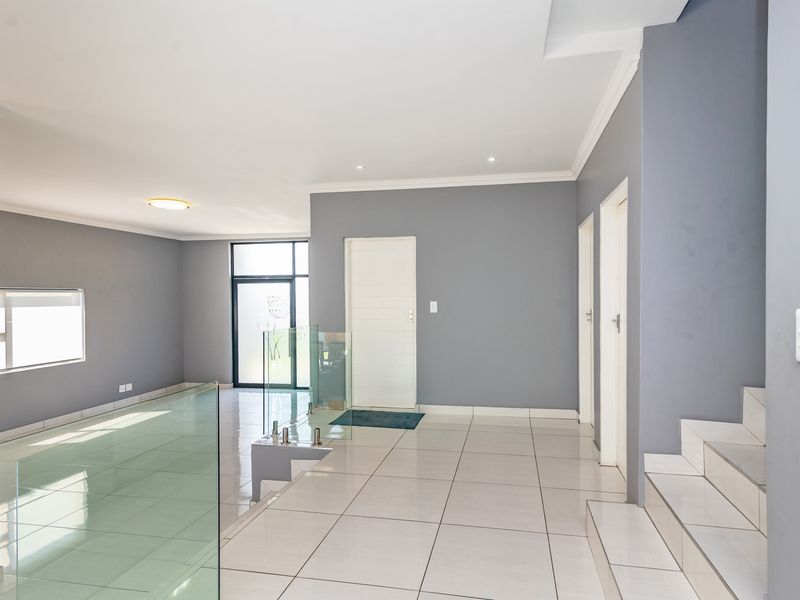 A spacious, modern hallway featuring neutral grey walls, white tiled flooring, and a contemporary glass balustrade overlooking the staircase.