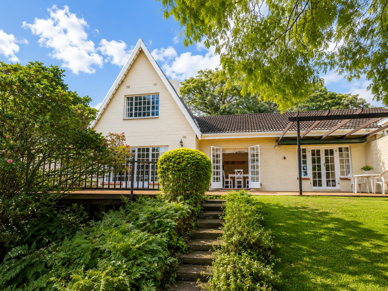 Rear view of a charming house featuring a timber deck, pergola, and well-maintained garden with concrete stairs leading to the main living area.