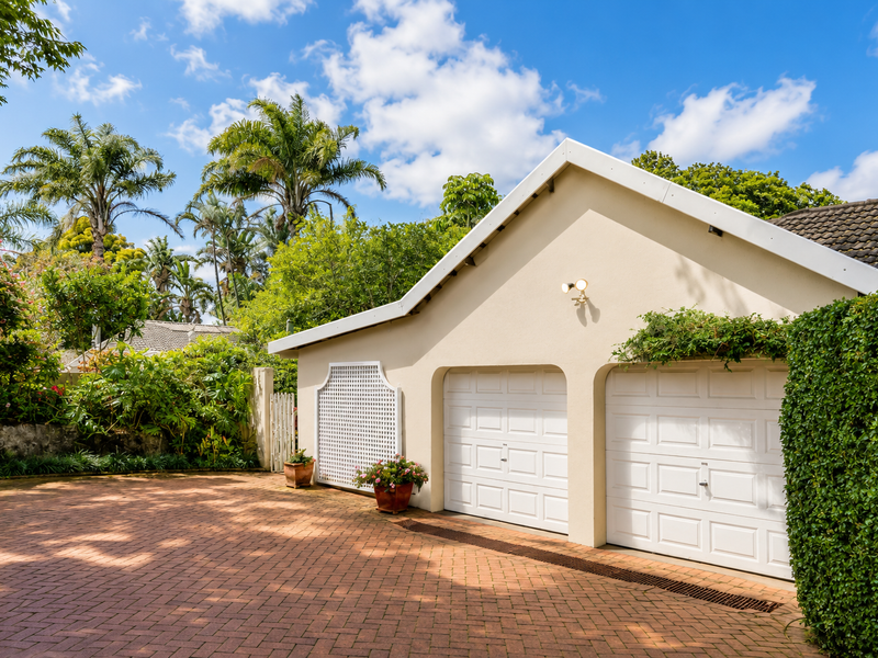 A well-maintained double garage with white panelled doors, set against a lush garden backdrop with a paved brick driveway.