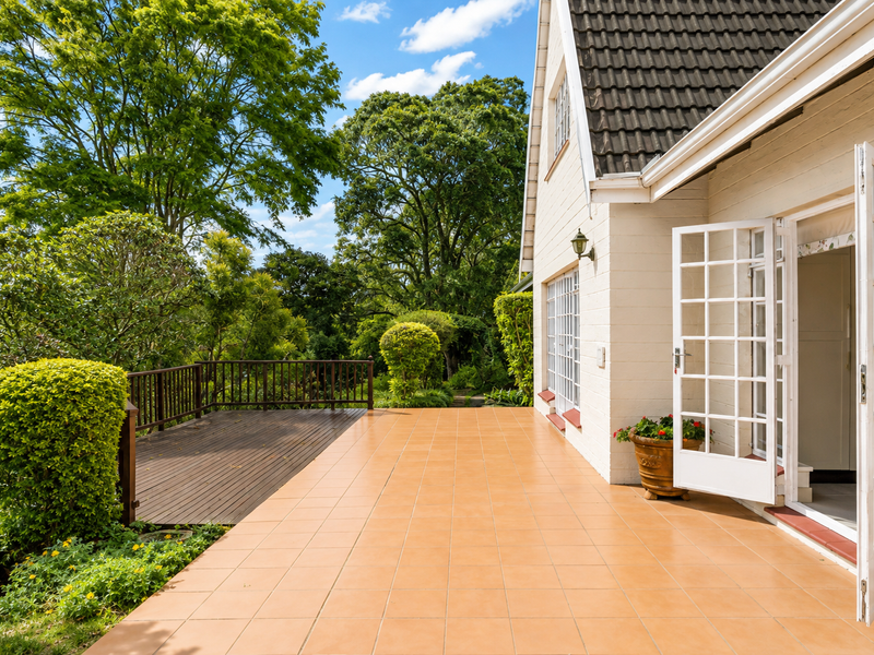 A spacious outdoor patio featuring terracotta-style tiling and an adjoining timber deck, accessible through classic white french doors, overlooking a lush garden.