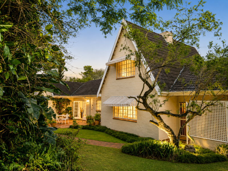 A charming rear exterior view of a house featuring a pitched tiled roof, brick facade, and a well-maintained garden with a paved walkway.