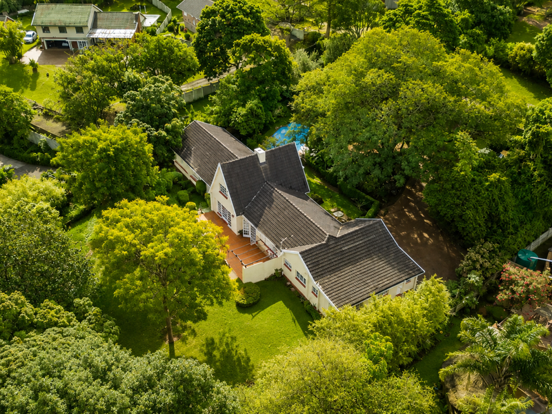 Aerial view of a residential property featuring a tiled roof house, a swimming pool, a paved patio area, and extensive mature garden landscaping.