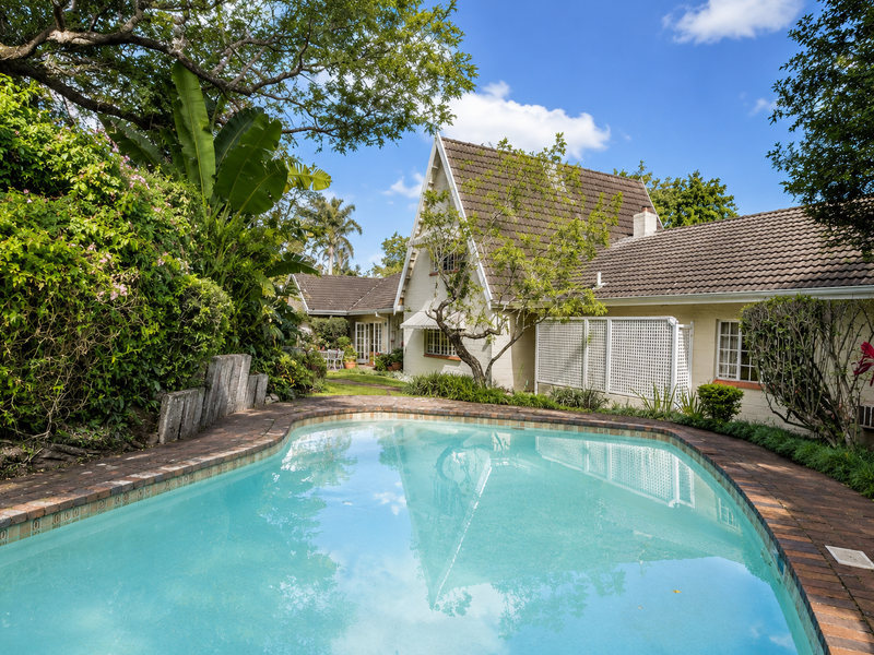 A well-maintained swimming pool set in a lush garden, featuring brick coping and a view of the charming pitched-roof house exterior.