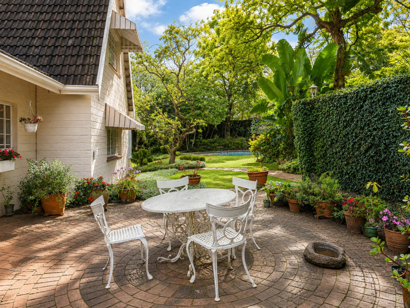 A charming rear garden featuring a circular brick-paved patio area, mature landscaping, and a well-maintained lawn leading toward a private pool area.