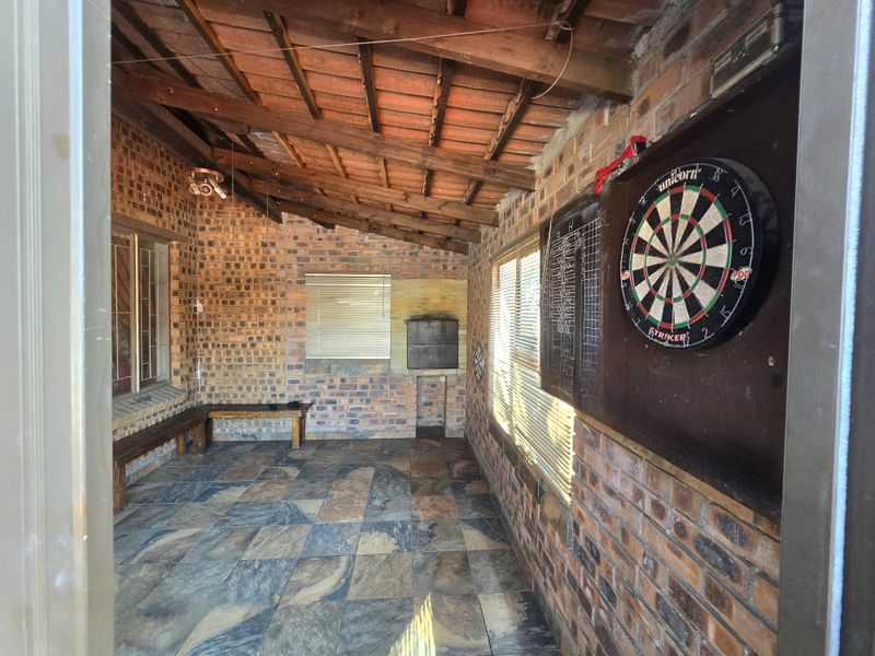 Covered outdoor patio featuring exposed brick walls, timber beam ceiling, tiled flooring, and a built-in braai area, ideal for entertainment.