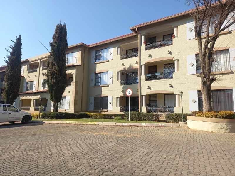 Exterior view of a multi-story apartment complex featuring a paved parking area, landscaped gardens, and private balconies with wrought iron railings.