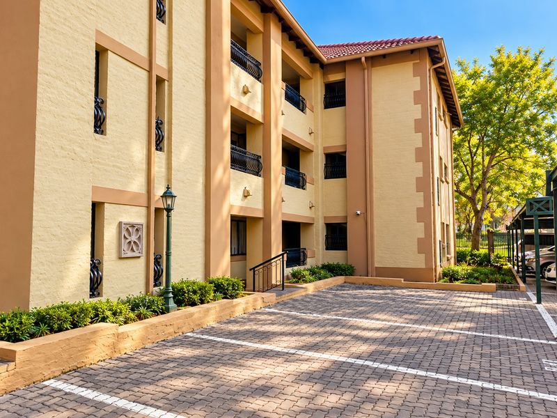 Exterior view of the apartment building facade featuring paved parking, wrought iron balcony railings, and well-maintained landscaped garden beds.