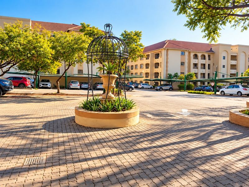 A well-maintained apartment complex exterior featuring paved parking areas, covered parking bays, and a central ornamental garden feature.
