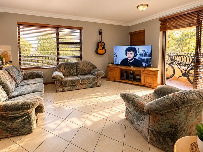 A bright living room featuring tiled flooring, crown molding, and large windows with venetian blinds, providing access to an outdoor balcony area.