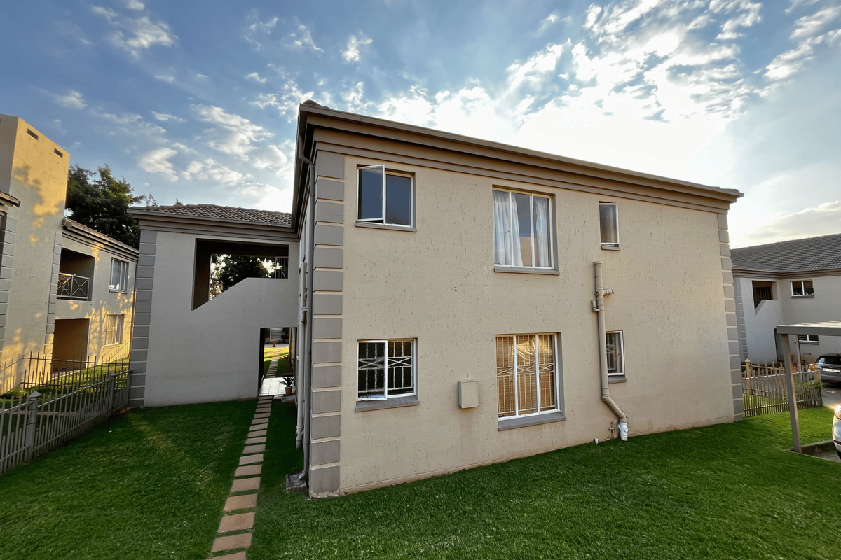 Rear view of a townhouse featuring a plastered facade, tiled roof, security bars on windows, and a well-maintained lawn with a paved walkway.