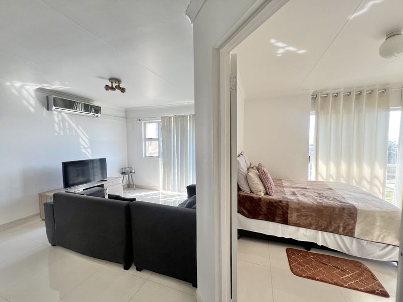 An open-plan living area adjacent to a bedroom, featuring tiled flooring, an air conditioning unit, and natural light through sliding doors.