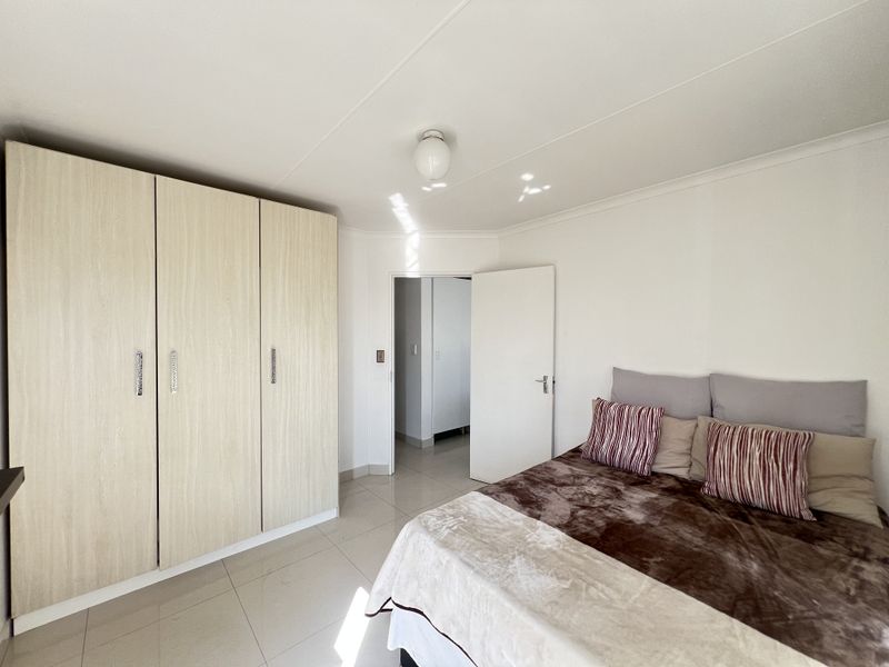 A bright bedroom featuring neutral tiled flooring, built-in wooden cupboards, and a clean, minimalist aesthetic with direct access to an adjacent hallway.