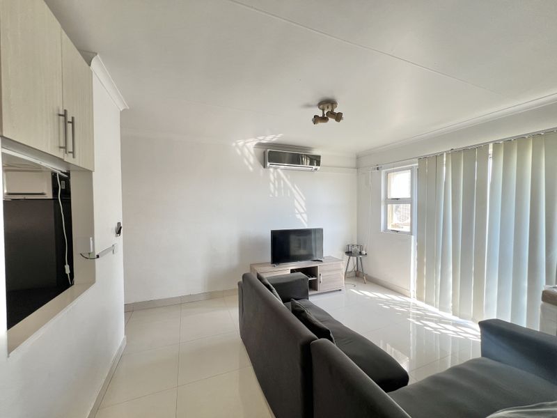 A bright living room featuring tiled flooring, a wall-mounted split-system air conditioner, and vertical blinds allowing natural light.