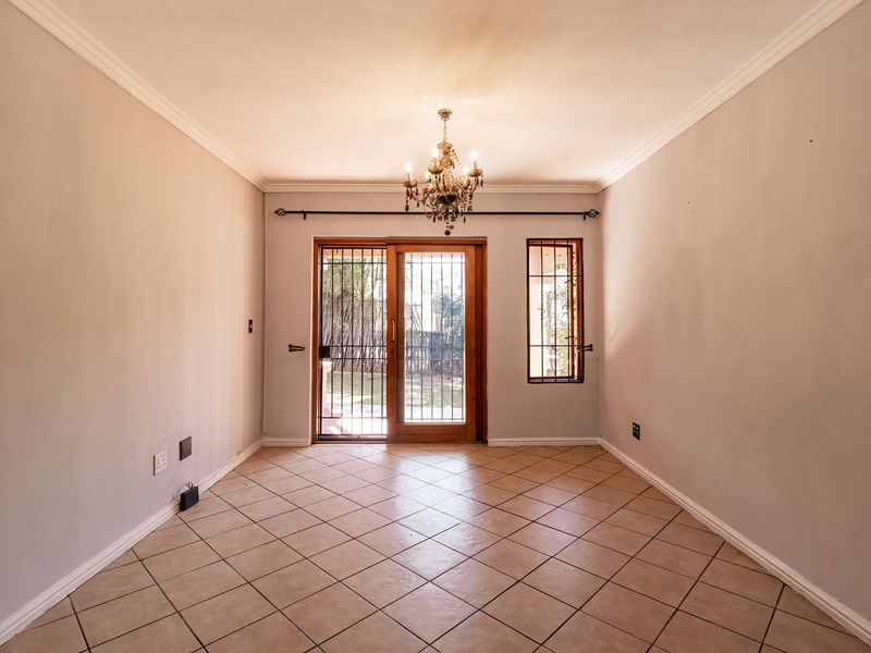 A bright, empty living room featuring tiled flooring, a decorative chandelier, and a sliding glass door with security gate leading to the garden.