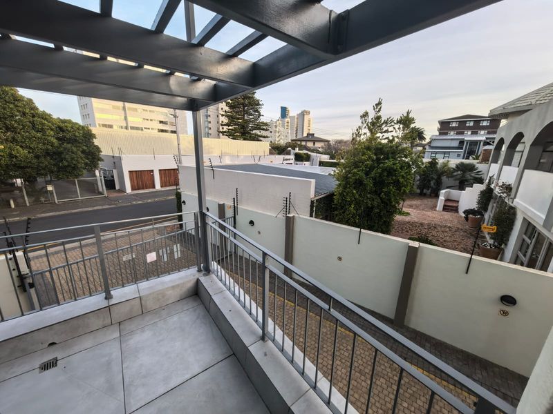 A modern balcony featuring a metal pergola, tiled flooring, and a metal balustrade, overlooking the street and surrounding residential area.