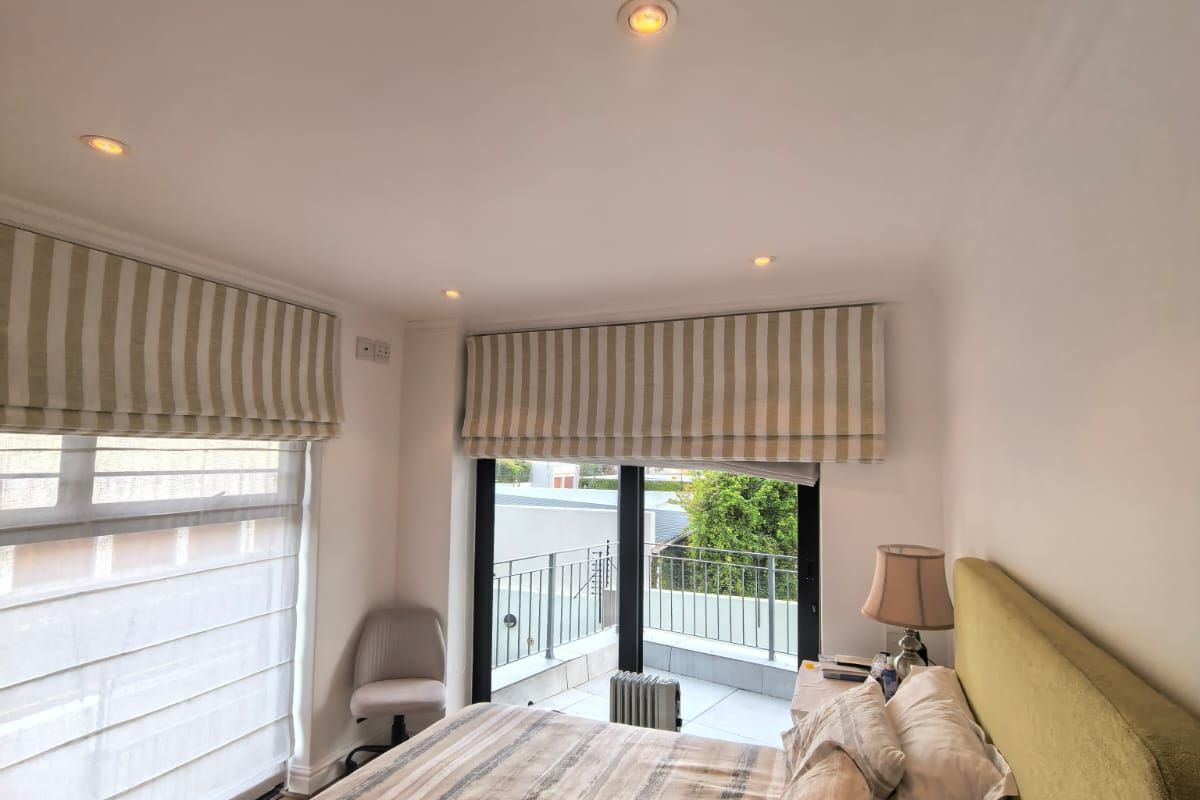 A bright and well-maintained bedroom featuring recessed lighting, large windows with roman blinds, and direct access to a private balcony.