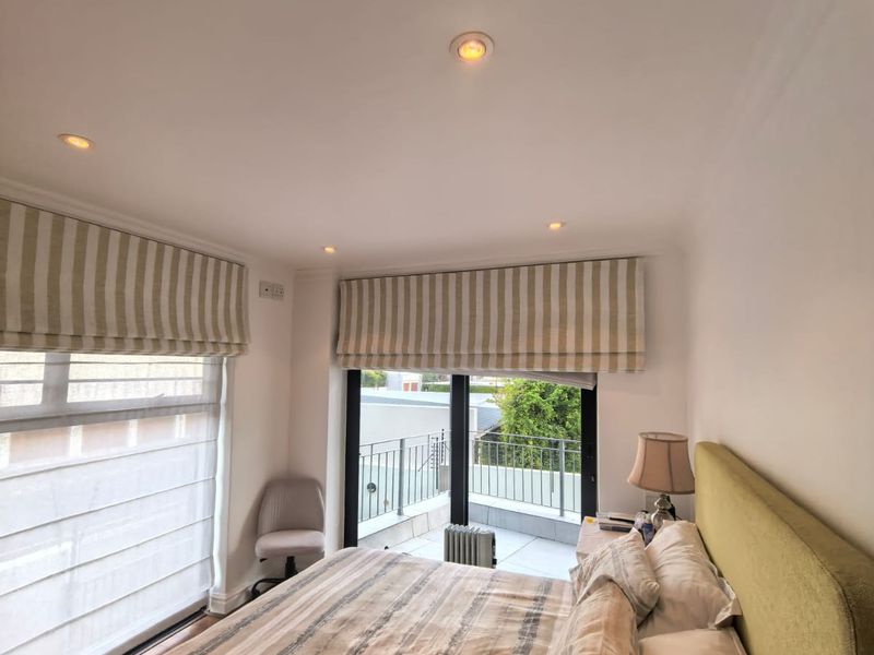 A bright and well-maintained bedroom featuring recessed lighting, large windows with roman blinds, and direct access to a private balcony.