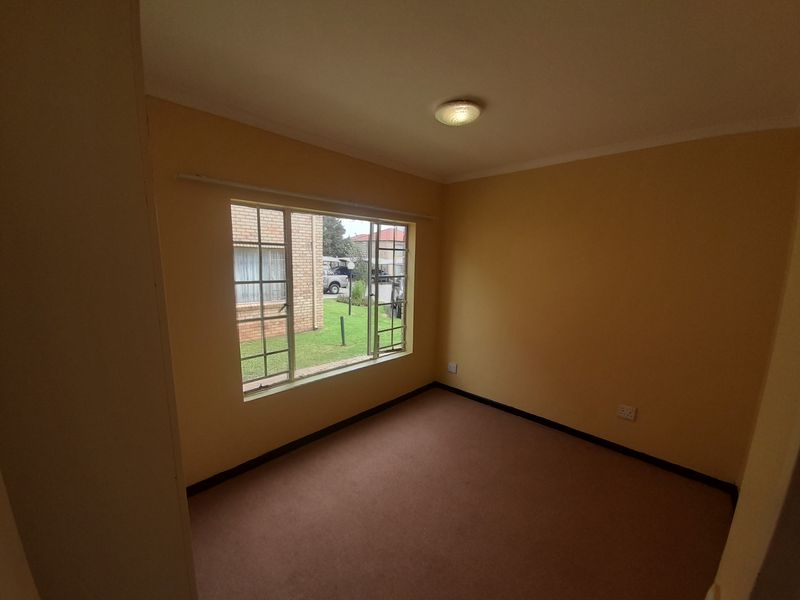 An empty bedroom featuring carpet flooring, neutral walls, and a large metal-framed window providing natural light and views of the exterior grounds.