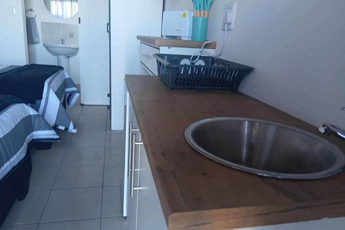 A compact kitchenette area featuring a wooden countertop, stainless steel sink, and built-in cabinetry, adjacent to a bedroom with a pedestal basin.