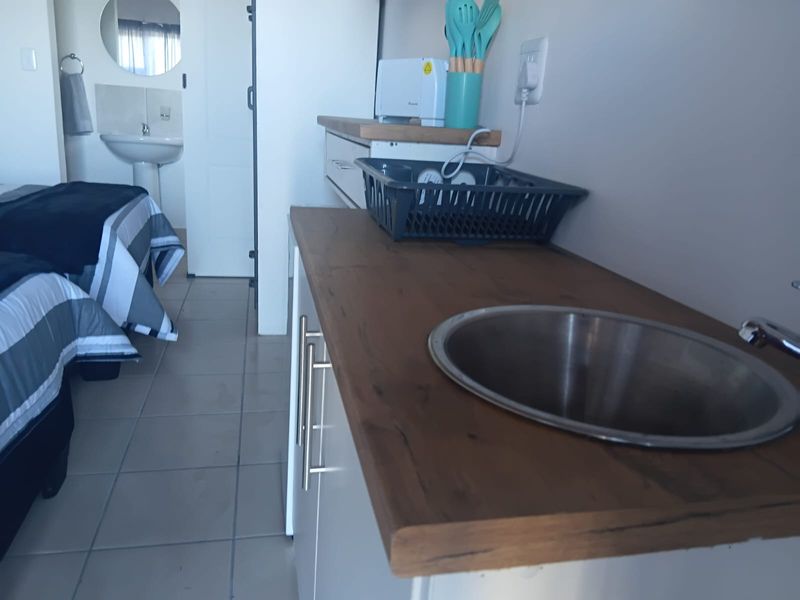 A compact kitchenette area featuring a wooden countertop, stainless steel sink, and built-in cabinetry, adjacent to a bedroom with a pedestal basin.