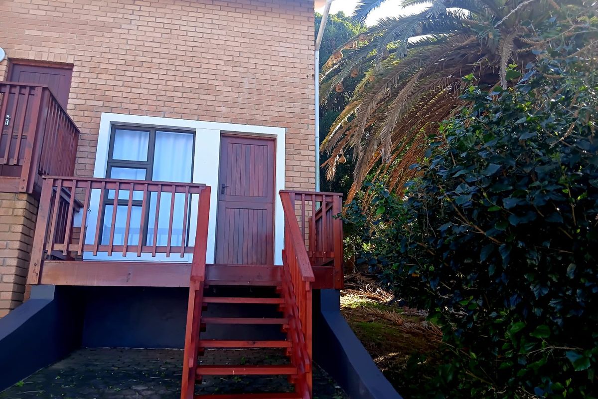 Rear exterior view of the cottage featuring a raised timber deck, staircase, and brick facade with access door and window.