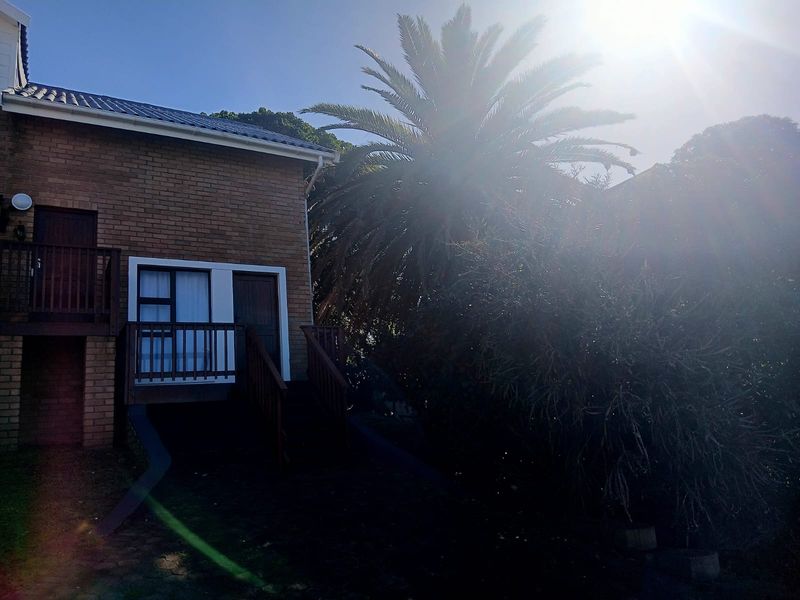 Rear view of a brick cottage featuring a timber balcony and staircase, set against a mature garden with significant lens flare.