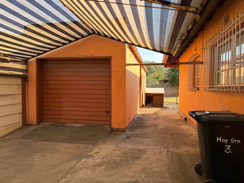 Exterior view of a garage with a concrete driveway and overhead shade cloth awning, featuring security bars on the adjacent building windows.