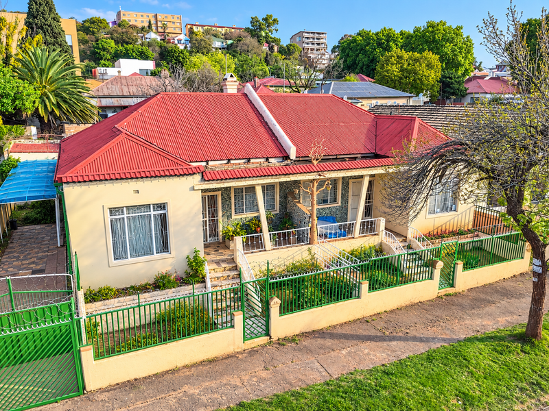 Front elevation of a residential house featuring a red corrugated metal roof, perimeter wall with security fencing, and a paved driveway.