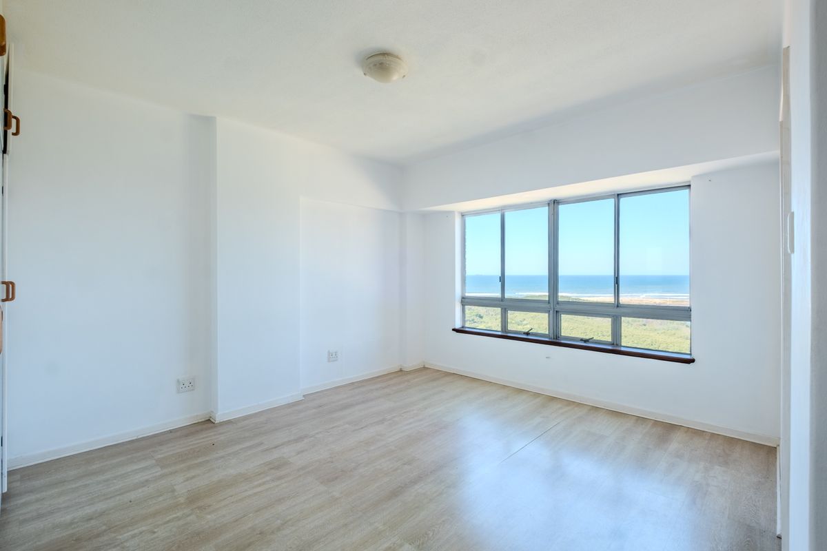 A bright, empty bedroom featuring light-toned laminate flooring and large windows offering a scenic ocean view.