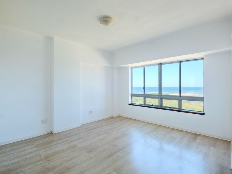 A bright, empty bedroom featuring light-toned laminate flooring and large windows offering a scenic ocean view.