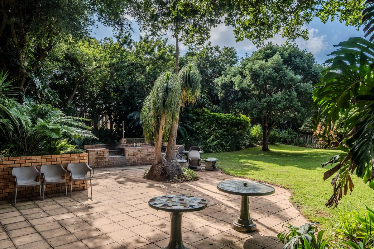 A spacious rear garden featuring a paved patio area with a built-in brick braai, surrounded by mature trees and established landscaping.