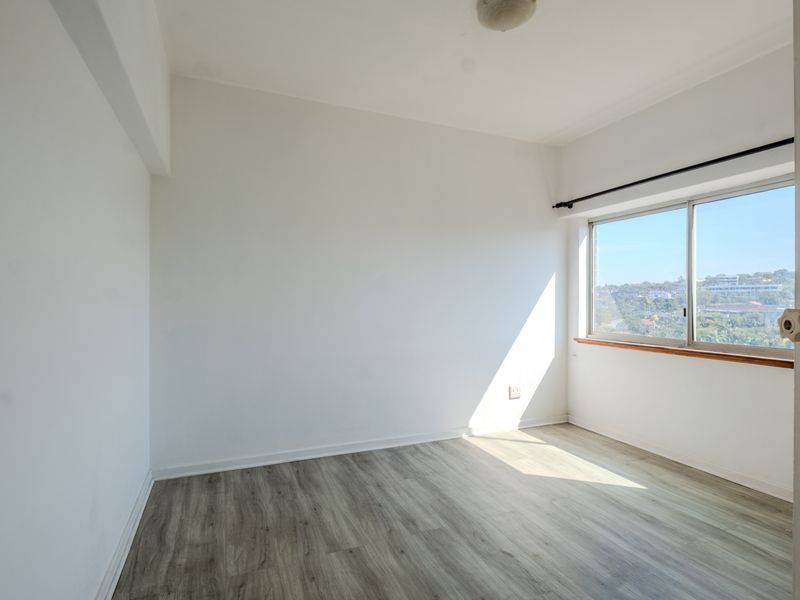 A bright, empty bedroom featuring modern laminate flooring, a large sliding window with natural light, and a neutral white finish.