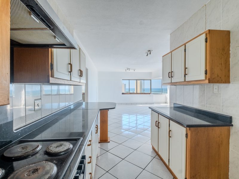 A functional kitchen featuring built-in cabinetry, granite countertops, and an electric hob, leading into an open-plan living area with ocean views.