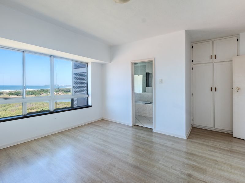 Bright and spacious bedroom featuring built-in cupboards, modern laminate flooring, and expansive windows offering scenic coastal views.