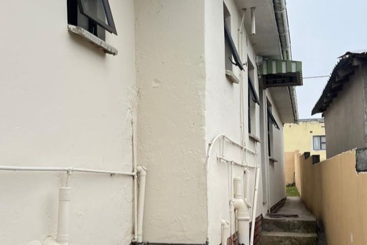 Side exterior view of the property showing exposed plumbing, brick foundation, and a concrete pathway leading to the rear.