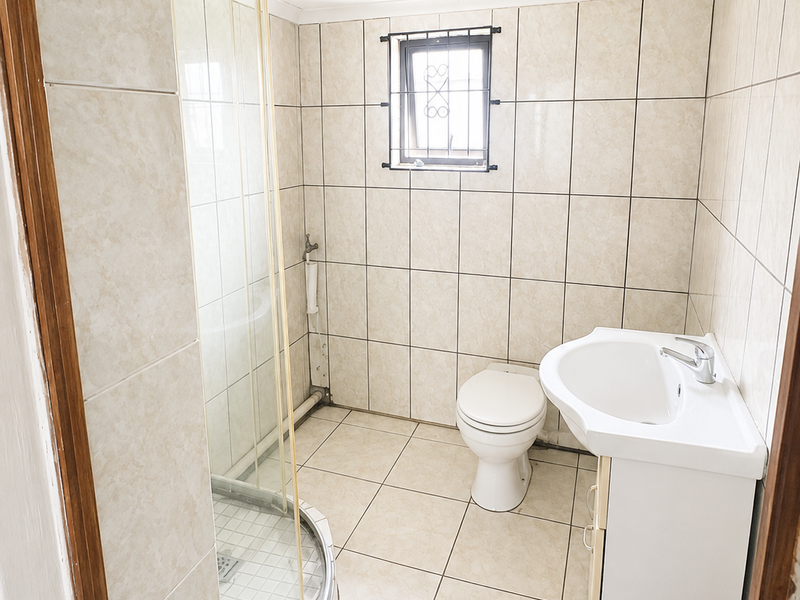 A functional bathroom featuring a corner shower enclosure, pedestal basin, and toilet, finished with neutral wall and floor tiling.