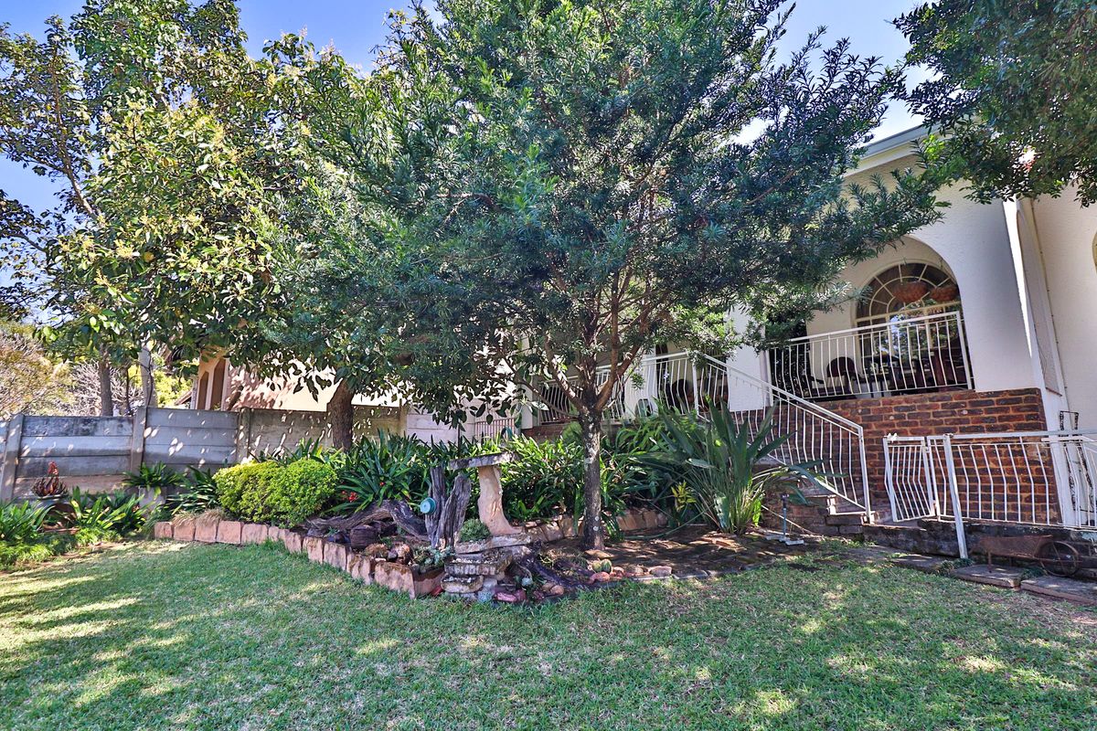 Rear garden view featuring a landscaped lawn, mature trees, brick retaining wall, and a raised patio area with metal balustrades and arched window detail.