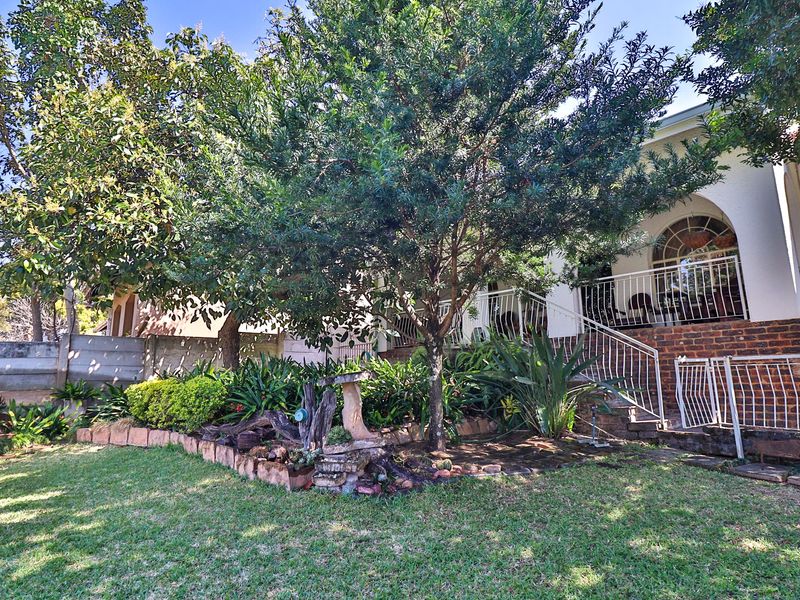 Rear garden view featuring a landscaped lawn, mature trees, brick retaining wall, and a raised patio area with metal balustrades and arched window detail.
