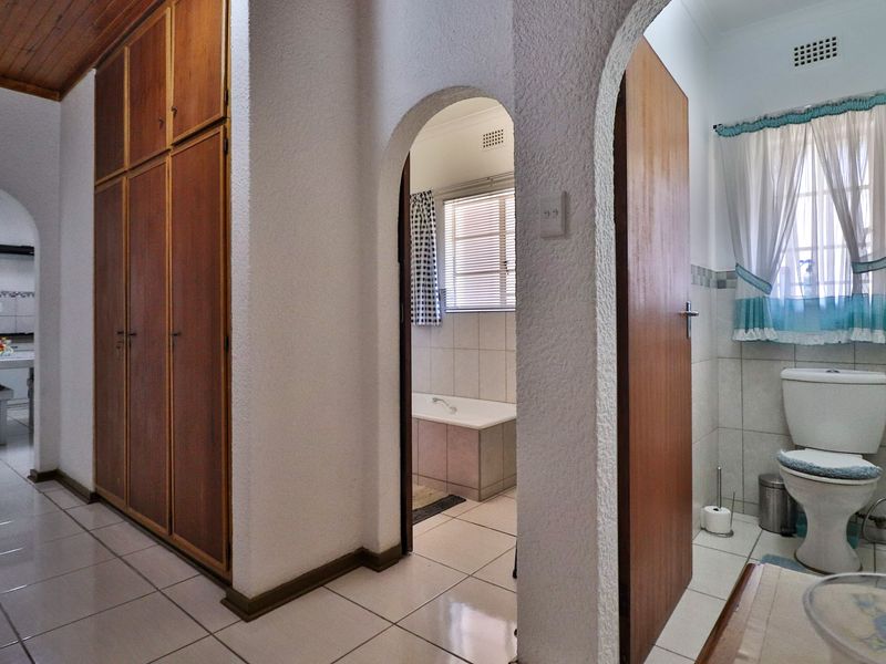 A hallway featuring built-in timber cupboards, tiled flooring, and arched entryways leading to a bathroom and kitchen area.