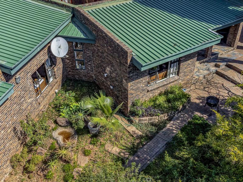 Aerial view of the rear exterior featuring a face brick facade, green corrugated metal roofing, and a paved patio area with established garden landscaping.
