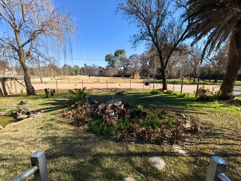 A spacious garden area featuring mature trees, established landscaping, and a wire fence boundary overlooking an open field.