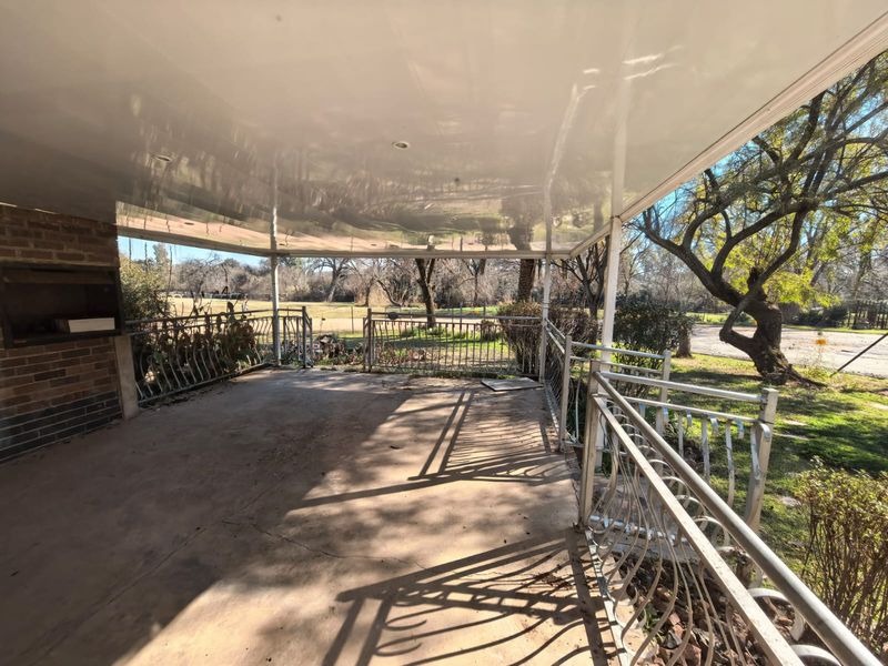 A spacious covered patio featuring a built-in braai and metal railings, overlooking a garden area with mature trees.