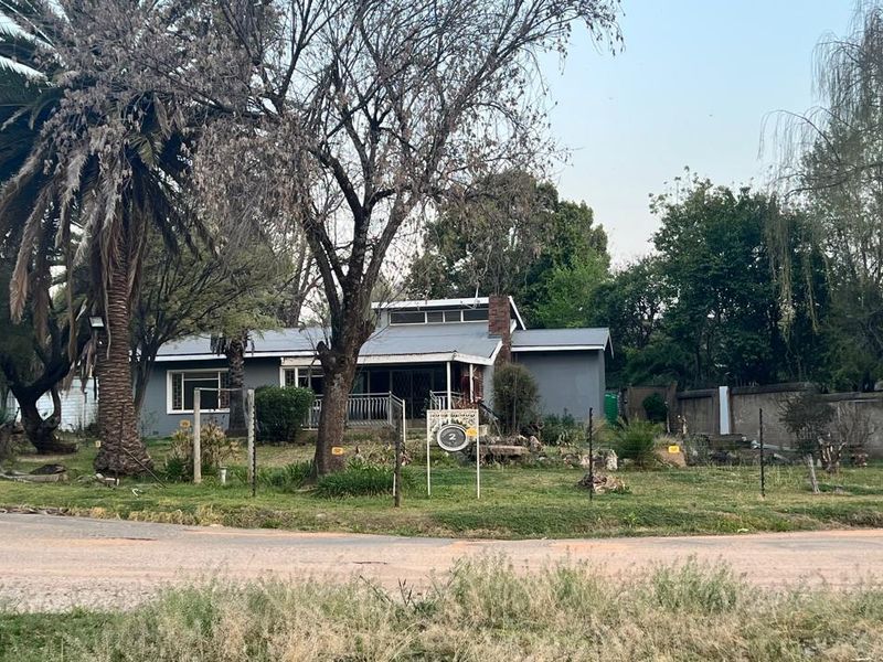 Front view of a single-story house featuring a metal roof, brick chimney, and a large, mature garden with perimeter fencing.